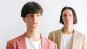 A imagem mostra dois homens representando o empreendedorismo LGBT+ O que está mais ao fundo é branco, cabelos castanhos, lisos e soltos em formato chanel, usa uma camisa branca e um paletó bege. O homem da frente é branco, tem cabelos curtos e castanhos, usa uma camisa branca e um paletó rosa.