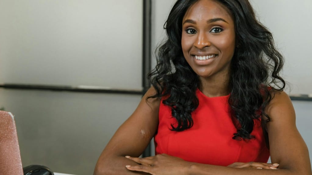 A imagem mostra uma mulher negra de cabelos ondulados, pretos e soltos. Ela está em uma sala, sentada a frente de um computador aberto, com os braços cruzados e apoiados em uma mesa. Ela olha para a câmera e sorri.