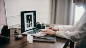 A imagem mostra apenas os braços e as mãos de um homem digitando em um computador que está apoiado em cima de uma mesa. Na tela do computador, uma foto de uma mulher sem termo de autorização de uso de imagem está sendo editada,