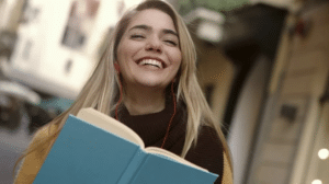 A imagem mostra uma jovem de cabelos loiros, longos e soltos que conseguiu acumular bolsa de pós-graduação. Ela está com uma blusa de frio de cor vinho e sorrindo. Nas mãos ela segura um caderno de capa dura azul.