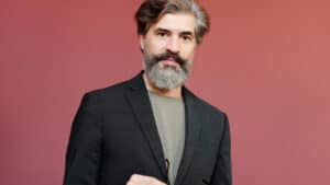 A imagem mostra um homem de cabelos e barba grisalhas. Ele usa uma blusa cinza e um blazer preto. Ele olha diretamente para a câmera em um fundo vermelho.