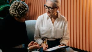 A imagem mostra duas mulheres sentadas em um sofá conversando em um ambiente de escritório. A mulher da direita faz parte da diretoria executiva. Ela é branca, tem cabelos brancos, lisos e presos em um rabo de cavalo, usa um óculos de grau e veste uma saia preta e uma blusa social branca de mangas compridas. Ela segura uma prancheta nas mãos enquanto olha para a mulher da direita. A mulher da direita é negra, tem cabelos enrolados e curtos, usa uma blusa social preta e conversa com a mulher da direita com o rosto virado para ela.