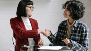 A imagem mostra duas mulheres conversando sobre MROSC. A mulher da esquerda é branca, de cabelos curtos, lisos e soltos até o ombro. Ela usa óculos de grau, uma blusa branca e um blazer bonina. A mulher da direita é branca, tem cabelos curtos, ondulados e soltos. Ela usa uma blusa rosa claro e um blusa xadrez de manga comprida por cima. As duas estão sentadas em uma mesa e olham uma para a outra enquanto apertam as mãos.
