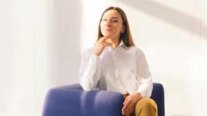 A imagem mostra uma mulher branca de cabelos marrons, lisos, longos e soltos, usando uma blusa social branca e uma calça de cor mostarda. Ela está em uma sala, sentada em uma poltrona azul, com a mão esquerda apoiada no queixo e um ar confiante pois sua ONG tem uma Consultoria Jurídica para o Terceiro Setor.