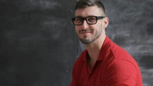 A imagem mostra um homem jovem branco de cabelos pretos e curtos e barba curta. Ele está em um local fechado com paredes pretas, usando óculos de grau e camisa social vermelha. Ele olha para a câmera com um olhar confiante devido ao reconhecimento da ausência de improbidade administrativa.