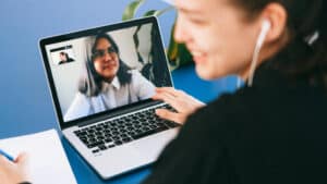 A imagem mostra duas mulheres em uma videochamada sobre controle social no Terceiro Setor. Uma delas aparece em primeiro plano, sorrindo enquanto olha para a tela de um notebook e segura uma caneta sobre um caderno com anotações. Na tela do computador, é possível ver a outra mulher, que usa óculos e uma camisa branca, também sorrindo durante a conversa. Ao fundo, há uma planta, e o cenário é iluminado por luz natural.