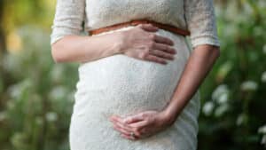 A imagem mostra uma a barriga de uma mulher grávida. Ela está em um ambiente aberto e com jardins, usa um vestido branco e coloca as mãos na barriga. Essa mulher vai poder desfrutar da nova lei da Licença-Maternidade Estendida.