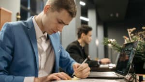 A imagem mostra um homem branco de cabelos loiros e curtos sentado em uma cadeira, em frente a uma mesa em um ambiente coletivo. Ele usa uma blusa social branca e um paletó azul. Ele está de cabeça baixa e olhando para um documento sobre responsabilidade pessoal de dirigentes enquanto escreve nele.