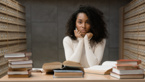 A imagem mostra uma mulher negra de cabelos pretos, crespos, longos e soltos. Ela está em um espaço que lembra a uma biblioteca, a frente de uma mesa cheia de livros. Ela usa uma blusa de manga comprida branca e está com os cotovelos apoiados na mesa e o rosto entre as mãos, com uma expressão de dúvida para saber se é possível acumular bolsa da fapesp com outra atividade remunerada.