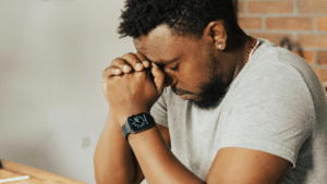 A imagem mostra um homem negro de cabelos crespos e curtos, usando uma blusa branca e um relógio de pulso. Ele está sentado, com as mãos apoiadas na testa e os olhos fechados, transmitindo um sentimento de preocupação e refletindo sobre a desistência da pós-graduação. Ele parece estar em uma sala com uma parede de tijolos laranjas e outra branca.