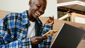 A imagem mostra uma mulher negra de cabelos curtos e loiros, usando uma blusa branca e uma camisa xadrez azul. Ela sorri enquanto olha para o celular e para o notebook ao mesmo tempo, buscando informações sobre como elaborar um plano de trabalho segundo o MROSC.