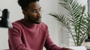 A imagem mostra um homem negro de cabelos curtos e crespos, usando um óculos de grau e uma blusa de mangas longas rosa. Ele está em um ambiente que parece ser um escritório bem iluminado. Ele está sentado a frente de uma mesa, com os olhos ficos em um computador, estudando quais são as despesas permitidas pelo MROSC.