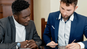 A imagem mostra dois homens sentados à mesa, em um ambiente interno. O homem da esquerda é negro, tem caeblos crespos, pretos e curtos, veste uma blusa branca e um paletó preto. O homem da direita é branco e tem cabelos lisos, marrons e curtos. Ele usa uma blusa branca e um paletó azul escuro. Eles estão inclinados sobre a mesa e o homem da direita assina um testamento.