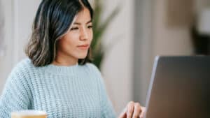 A imagem mostra uma mulher branca de cabelos pretos, lisos e na altura dos ombros. Ela está sentada à mesa, usando um suéter azul claro, enquanto trabalha em um notebook. Ela está em um ambiente iluminado e parece concentrada nos estudos sobre reembolso e remuneração no trabalho voluntário.