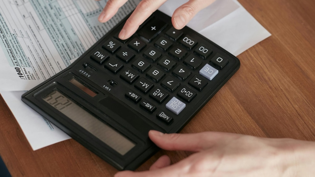 A imagem mostra mãos de uma pessoa branca utilizando uma calculadora sobre uma mesa de madeira, ao lado de documentos e formulários financeiros. A cena remete à análise contábil e organização sobre tributação de doações no Terceiro Setor.