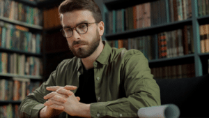 A imagem mostra um homem branco de cabelo castanho e barba curta. Ele usa óculos de grau, uma blusa preta e um blusa social verde escurto de mangas longas por cima, Ele está sentado à mesa em uma biblioteca, com várias estantes de livros ao fundo. As mãos estão entrelaçadas sobre a mesa e ele tem um olhar concentrado, enquanto reflete se bolsista no exterior pode responder por peculato.
