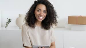 A imagem mostra uma mulher de pele negra, cabelos crespos, longos e solos, usando uma blusa de mangas curtas de cor bege. Ela está sorrindo em um ambiente iluminado, com um tablet na mão com um texto sobre o papel do terceiro setor na promoção de Direitos Humanos.