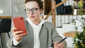 Uma mulher branca de cabelos ruivos e presos em uma trança está sentada à mesa em um ambiente interno, segurando um celular vermelho com a mão esquerda e uma caneta com a mão direita, como se estivesse lendo algo na tela enquanto faz anotações sobre como pejotização no Terceiro Setor. Ela usa óculos de armação escura, blusa clara com capuz e um blazer cinza.