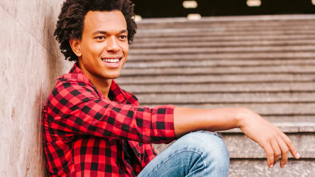 A imagem mostra um homem negro com cabelos curtos e enrolados, sentado em uma escada de pedra, apoiando as costas em uma parede. Ele usa uma camisa xadrez vermelha e preta e calça jeans clara, com um dos braços apoiado sobre o joelho dobrado. Seu rosto está voltado para o lado, com expressão sorridente pois acaba de tirar suas dúvidas sobre cobrança de bolsistas que estudaram no exterior com um professor.
