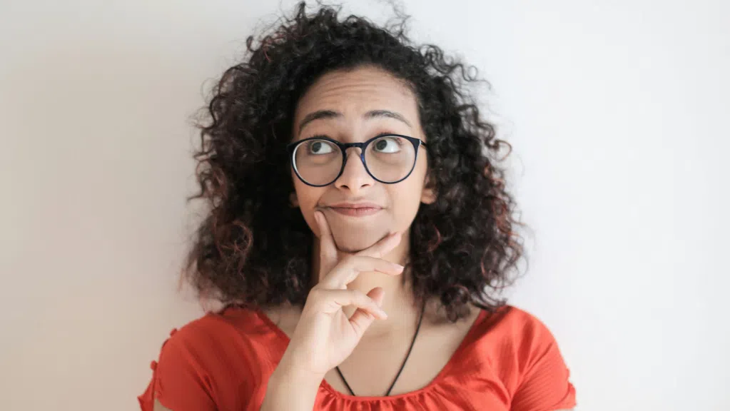 Uma mulher com cabelo cacheado e volumoso aparece em frente a um fundo claro, usando uma blusa laranja e óculos de armação escura. Ela está com a mão apoiada no queixo, olhando para cima, com expressão pensativa sobre negociar dívida com Capes ou CNPq.