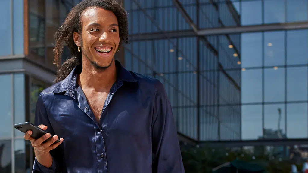 Uma pessoa com cabelos longos e cacheados está ao ar livre, sorrindo e olhando para o lado enquanto segura um celular na mão, vestindo uma camisa azul escura de manga longa. Ao fundo há um prédio moderno com fachada de vidro e estruturas metálicas refletindo a luz do dia. A pessoa está corrindo e segurando um celular na mão, enquanto pesquisa sobre o uso do nome social em concursos publicos.