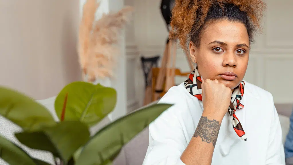 A imagem mostra uma mulher negra com cabelos crespos e presos em um coque alto. Ela está sentada, apoiando o queixo na mão, olhando diretamente para frente. Ela veste uma camisa branca e um lenço estampado no pescoço. No antebraço, há uma tatuagem. À frente, parcialmente desfocada, aparece uma planta com folhas verdes, e ao fundo há móveis e objetos de um ambiente interno. O tema parcerias sem chamamento público está associado ao conteúdo.