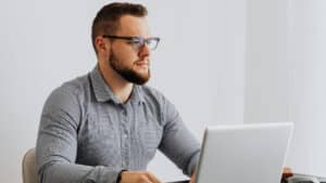Um homem branco, de cabelos loiros e curtos, barba curta e óculos de grau, está sentado à mesa usando um notebook. Ele veste camisa social cinza e olha atentamente para a tela, em um ambiente claro e neutro. A imagem remete a contexto profissional, como análise de documentos relacionados à Tomada de Contas Especial no MROSC.