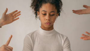 A imagem mostra uma mulher negra, jovem, com cabelo cacheado preso em um rabo. Ela usa uma blusa de gola alta branca. A mulher aparece em primeiro plano, olhando para baixo, com expressão triste por estar sofrendo assedio moral no trabalho. Ao redor dela, quatro mãos apontam em sua direção, vindas das laterais da imagem.
