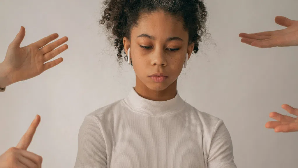 A imagem mostra uma mulher negra, jovem, com cabelo cacheado preso em um rabo. Ela usa uma blusa de gola alta branca. A mulher aparece em primeiro plano, olhando para baixo, com expressão triste por estar sofrendo assedio moral no trabalho. Ao redor dela, quatro mãos apontam em sua direção, vindas das laterais da imagem.