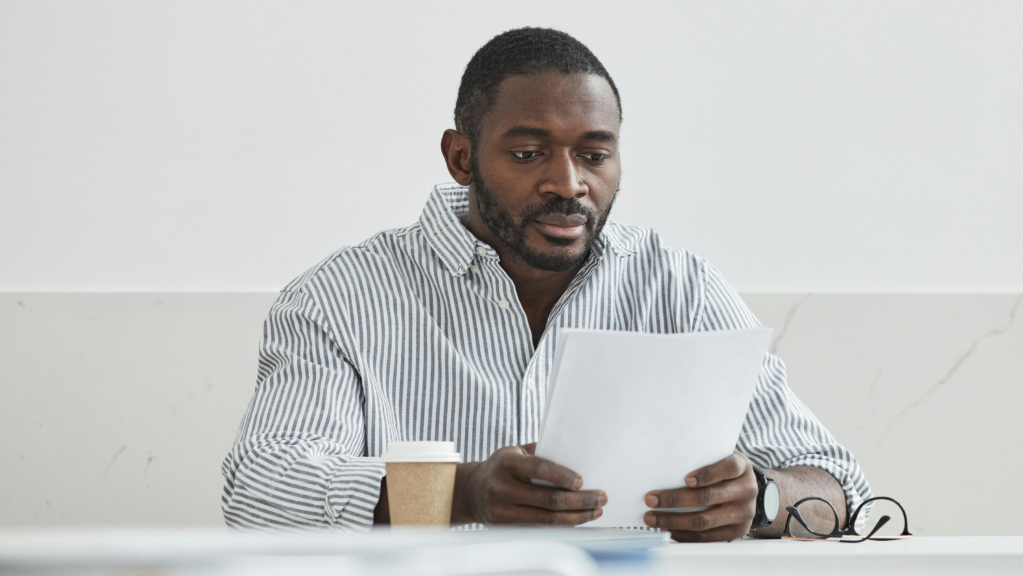 A imagem mostra um homem negro de cabelos pretos e curtos e barba completa. Ele usa uma blusa social de manga longa, listrada de azul e branco. O homem está sentado em uma cadeira, suas mãos seguram um papel enquanto estão apoiadas em uma mesa. Ele lê com atenção o documento sobre captação de recursos via emenda parlamentar.