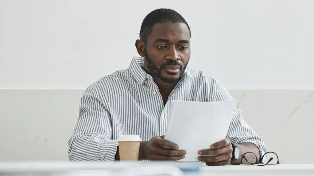 A imagem mostra um homem negro de cabelos pretos e curtos e barba completa. Ele usa uma blusa social de manga longa, listrada de azul e branco. O homem está sentado em uma cadeira, suas mãos seguram um papel enquanto estão apoiadas em uma mesa. Ele lê com atenção o documento sobre captação de recursos via emenda parlamentar.