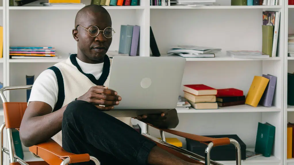 A imagem mostra um homem negro sentado em uma cadeira com braços de madeira e estrutura metálica. O homem usa óculos de grau, camiseta branca e um colete claro. Ele segura um notebook apoiado no colo e olha para a tela enquanto lê um conteúdo sobre chamamento público no MROSC. Ao fundo, há uma estante branca com livros, além de alguns objetos decorativos.