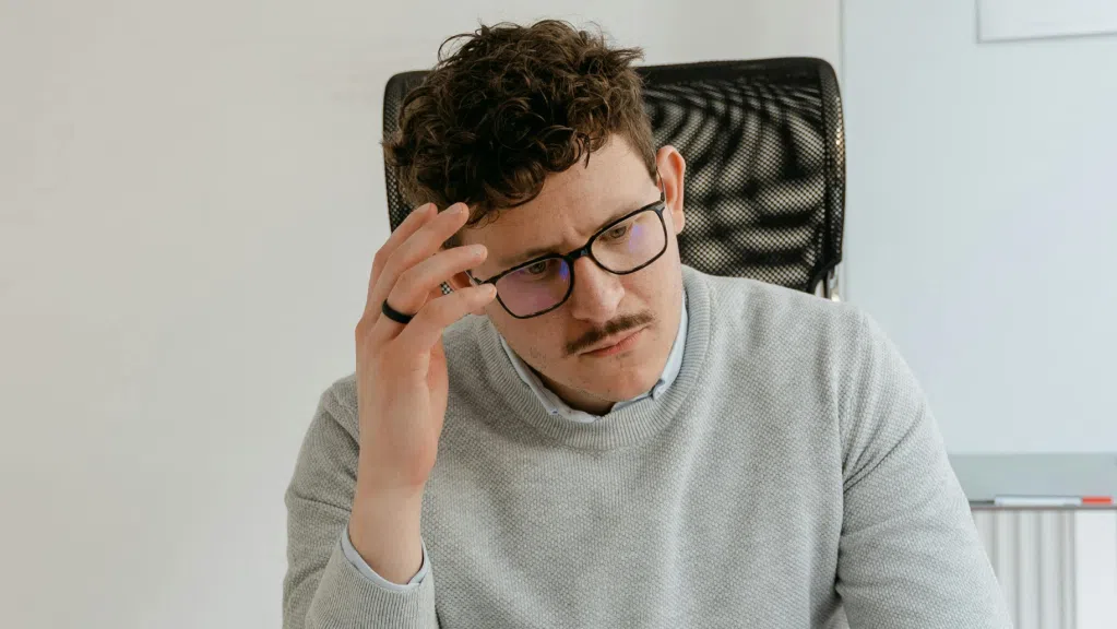 A imagem mostra um homem branco de cabelos curtos e bigode. Ele usa um óculos de grau e um moletom de cor cinza. Ele está sentado em uma cadeira de escritório, em um ambiente iluminado e leva a mão direita à cabeça com ares de preocupação pois precisa comprovar preterição em concurso público.