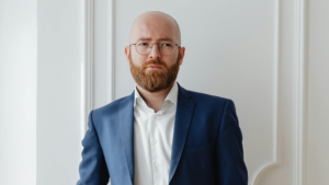A imagem mostra um homem branco, careca com barba ruiva e cheia. Ele usa óculos de armação fina e redonda e veste uma camisa de botões branca e um blazer azul escuro. O fundo é claro, com uma parede branca e molduras decorativas. O homem está com uma expressão confusa, tentando compreender as consequências da responsabilização do servidor público.