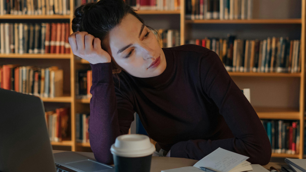 A imagem mostra uma mulher jovem e branca, de cabelos pretos e presos em um rabo, usando uma blusa de frio azul. Ela está em uma biblioteca (tem uma estante cheia de livros atrás) e sentada em uma cadeira, com um laptop aberto em uma mesa a sua frente. Ela está com a cabeça apoiada em uma das mãos enquanto olha para o lado com um olhar desolador pensando sobre a sua desistência de bolsa de estudos no exterior.