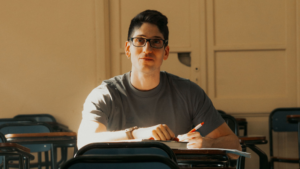 A imagem mostra um homem branco jovem de cabelos curtos e pretos, usando um óculos de grau e uma blusa de frio verde escura. Ele está em um ambiente fechado com uma caneta na mão estudando sobre o que fazer após ter sido aprovado fora das vagas de um edital de concurso público.