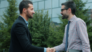 A imagem mostra dois homens brancos de cabelos curtos de cor marrom e barba. O home da esqueda usa uma blusa azul e um terno azul marinho. O homem da esquerda usa uma blusa social listrada e uma gravata azul escura. Eles estão se olhando nos olhos e apertando as mãos: são atores do MROSC e acabaram de firma uma parceria.