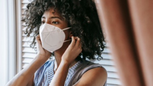 A imagem mostra uma mulher negra de cabelos crespos, longos e soltos. Ela está em um quarto, de costas para uma janela. Ela usa uma blusa azul e leva às mãos até as orelhas para colocar uma máscara de proteção individual enquanto batalha contra uma dispensa discriminatória por doença grave.