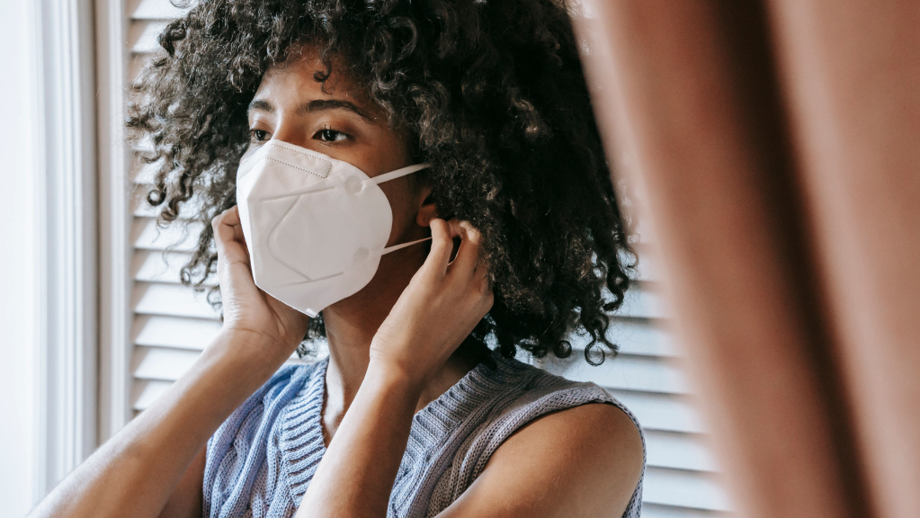 A imagem mostra uma mulher negra de cabelos crespos, longos e soltos. Ela está em um quarto, de costas para uma janela. Ela usa uma blusa azul e leva às mãos até as orelhas para colocar uma máscara de proteção individual enquanto batalha contra uma dispensa discriminatória por doença grave.