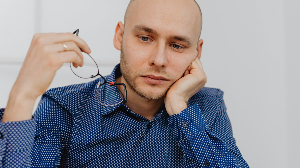 A imagem mostra um homem branco, careca de barba curta, usando uma blusa social azul. Ele está em um ambiente bem iluminado, com uma parede branca ao fundo. Ele segura o rosto com uma das mãos enquanto olha para baixo com um olhar triste enquanto reflete sobre as penalidades trabalhistas enfrentadas pela sua empresa.