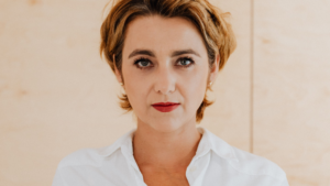 A imagem mostra uma mulher branca de cabelos curtos, loiros e soltos. Ela usa maquiagem e uma blusa social branca. Ela está em um ambiente iluminado, a frente de uma parede rosa clara. Ela olha diretamente para a câmera com um olhar confiante a respeito da Portaria Capes 180/2026.