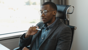 A imagem mostra um homem negro de cabelos curtos e óculos de grau. Ele usa uma camisa azul e um terno preto. Está sentado em uma cadeira de encosto preto, dentro de um escritório de paredes brancas. Ele segura uma caneta em uma das mãos e reflete sobre a reforma administrativa.