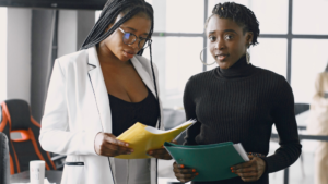A imagem mostra duas mulheres em um escritório bem iluminado. A mulher da esquerda é negra, tem cabelos longos e trançados, está usando um óculos de grau, blusa preta e blazer branco. A mulher da direita é negra, tem cabelo curtos, crespos e presos, e está usando uma blusa de manga comprida preta. A mulher da esquerda está olhando para uma pasta amarela que segura nas mãos sobre como a linguagem pode gerar vínculo empregatício enquanto a mulher da diretia olha diretamente para a câmera.