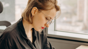 A imagem mostra uma mulher branca de cabelos curtos, louros e lisos. Ela usa óculos escuros, batom vermelho e uma blusa social preta. Ela está em um escritório e olhar para baixo enquanto analisa um papel sobre improbidade administrativa que está nas suas mãos.