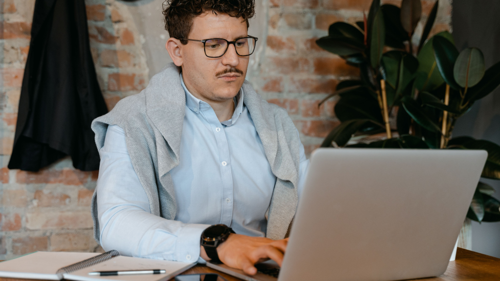 A imagem mostra um homem branco de cabelos enrolados e curtos, bigode e óculos de grau. Ele está em um escritório, com parede ao fundo de tijolo aparente. O homem usa uma blusa social azul clara e um casaco de lã cinza jogado nas costas. Ele está sentado a frente de uma mesa teclando em um computador aberto onde faz a seguinte questão: OSCIP ajuda a captar recursos?