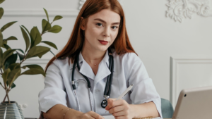 A imagem mostra uma mulher branca de cabelos longos, soltos e ruivos. Ela está em um consultório iluminado, de paredes brancas, sentada em uma cadeira em frente a uma mesa, buscando informações sobre Auxílio-moradia para médico residente. Ela usa um jaleco branco e um estetoscópio pendurado no pescoço.