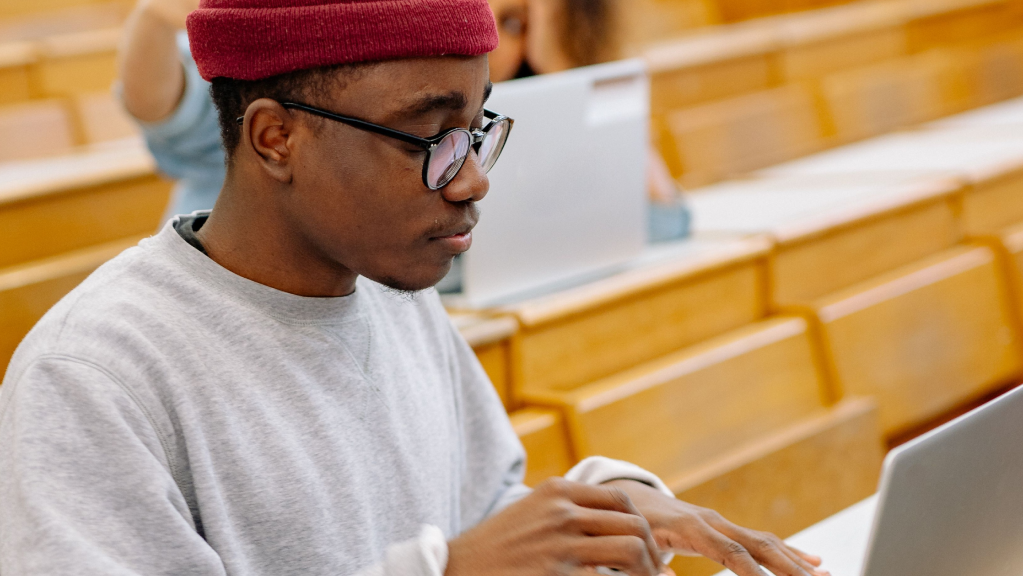 A imagem mostra um homem negro usando um gorro de lã vermelho e um óculos de grau. Ele está em um ambiente que parece um auditório bem iluminado, com cadeiras de madeira clara. Ele está sentado em uma delas e digita em um computador enquanto pesquisa sobre dolo, culpa ou boa-fé.