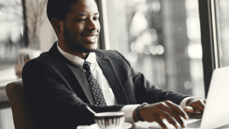A imagem mostra um homem negro de cabelos crespos e curtos. Ele usa uma blusa social branca e um terno preto com gravata preta. Ele está sentado em um escritório bem iluminado, usando um computador e com um sorriso pois acaba de recber a aprovação do seu afastamento para estudos.