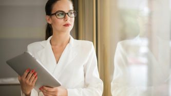 Uma pessoa está em pé, próxima a uma janela, segurando um tablet com as duas mãos onde lê um conteúdo sobre benefícios para PJ. Essa pessoa é uma mulher branca de cabelos castanhos, lisos e presos em um rabo de cavalo. Ela usa óculos e veste uma blusa branca de manga longa. Seu olhar está direcionado para fora.