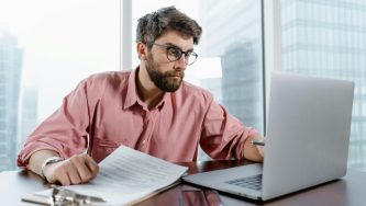 A imagem mostra um homem com cabelos e barba grisalha fazendo inscrição no CNU 2025. Ele usa um óculos de grau e uma blusa social de manga comprida rosa. O homem está sentado em uma cadeira e tem, à sua frente, um computador apoiado em uma mesa. Ele está olhando para o computador e mexendo no teclado com a mão esquerda, enquanto anota algo em um papel branco com a mão direita.